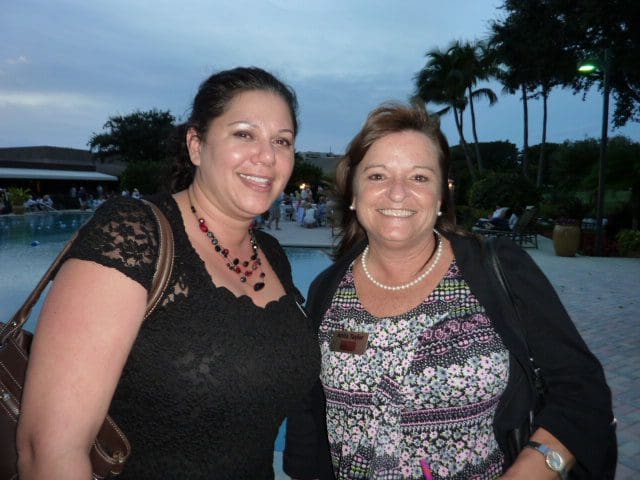 Two women smiling outdoors near water and palm trees at dusk.